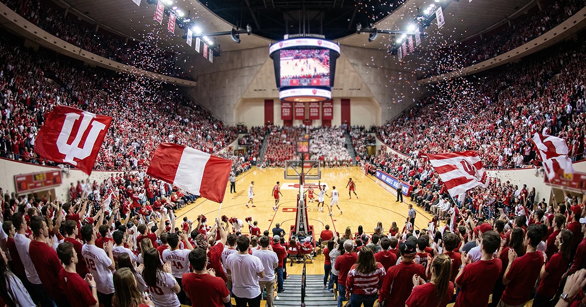 Pabellón universitario de baloncesto lleno de aficionados con banderas y colores de equipos